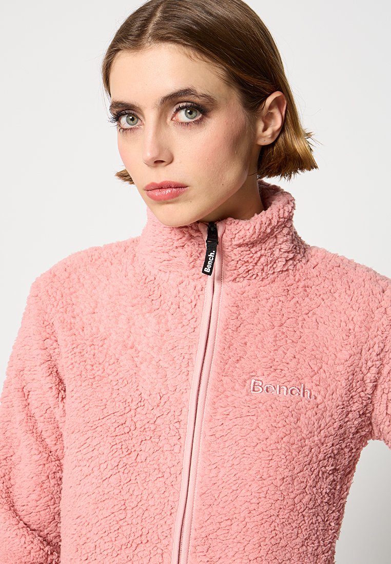 Różowa polarowa kurtka z miękkim, teksturowanym wykończeniem, wysokim kołnierzem i zamkiem błyskawicznym. Posiada tonalne, haftowane logo na piersi.