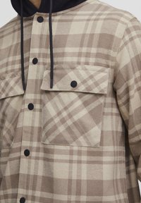 Geruite flanellen shirt in beige en bruin, met een knoopsluiting aan de voorkant, twee borstzakken met kleppen en een zwarte trekkoordcapuchon.
