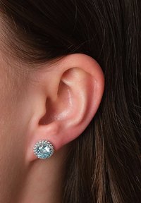 Nahaufnahme eines Ohrs mit einem silbernen Steckerohrring, der einen großen hellblauen Edelstein zeigt, der von kleinen klaren Steinen umgeben ist.