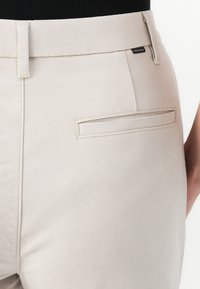 Beige cotton pants with a smooth texture, featuring a flat back pocket, belt loops, and a discreet brand label on the waistband.