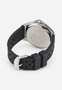 Reloj Timex con caja redonda de acero inoxidable, fondo transparente y correa de goma negra con perforaciones y hebilla plateada.