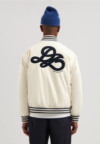 Hvid bomberjakke med marineblå stribede manchetter og kant, med en stor navy broderet logo på ryggen. Modellen bærer en blå strikket hue.