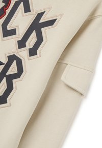 Beige sweatshirt met gedurfde geborduurde letters in zwart en rood. Zachte stof met geribbelde manchetten aan één mouw.