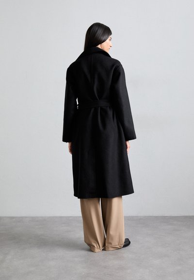 Μαύρο παλτό από μαλλί με μέση που ζώνει, μακριά μανίκια και φαρδύ γιακά. Το μοντέλο φοράει μπεζ παντελόνι σε φαρδιά γραμμή και μαύρες παπούτσια.