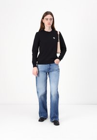 Felpa nera con scollatura rotonda e logo, abbinata a jeans chiari a gamba larga. La modella tiene in mano una borsa di colore chiaro.