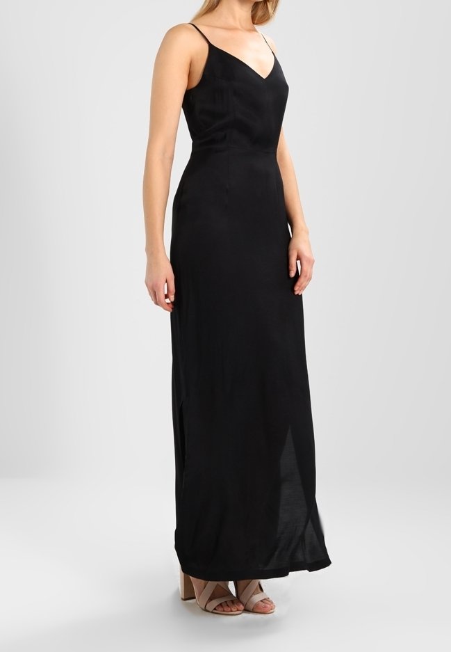 Robe maxi noire sans manches avec un décolleté en V ; tissu léger ; fentes sur les côtés ; le modèle porte des sandales à talons bloc couleur nude.