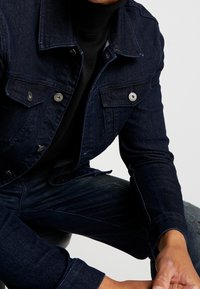 Chaqueta de mezclilla azul oscuro con dos bolsillos en el pecho, botones de presión plateados y un diseño ajustado. Combinada con un suéter negro de cuello alto y jeans desgastados.