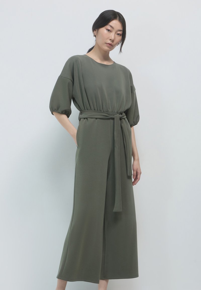 Giubbotto verde oliva realizzato in tessuto morbido, con scollatura rotonda, maniche corte a sbuffo e una cintura coordinata in vita.