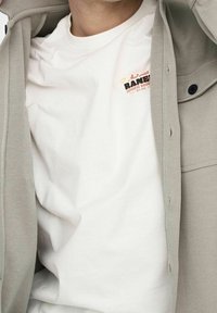 White cotton t-shirt with a front graphic featuring "Ramen" text in red and orange, worn under a gray, buttoned outer shirt.