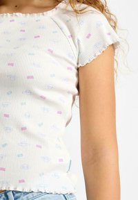 Hvid ribbet top med pastelfarvede mønstre af former. Har en bølget krave og korte ærmer, med en tætsiddende silhuet.