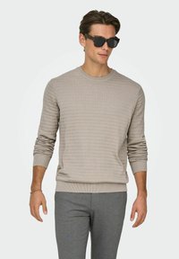 Pull en maille beige avec un motif texturé, col rond et poignets côtelés, associé à un pantalon gris ajusté et des lunettes de soleil foncées.