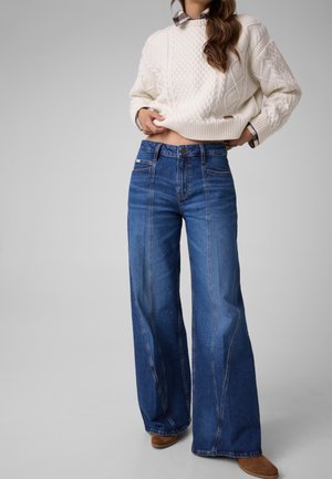 Kvinde iført blå jeans med vide ben, en hvid strikket sweater over en ternet skjorte og brune sko mod en ensfarvet lys grå baggrund.