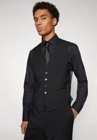 Schwarze maßgeschneiderte Weste mit schlankem Design, ausgestattet mit fünf Knöpfen, seitlichen Taschen und strukturiertem Stoff, kombiniert mit einer passenden Krawatte und Hemd.
