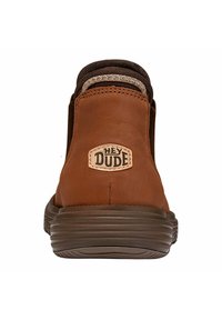Botas de cuero marrón hasta el tobillo con paneles de tela texturizada, puntera redonda y suela de goma. Presentan un logo "Hey Dude" en una etiqueta bordada.