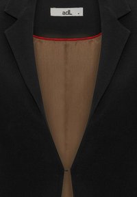 Blazer nero con rever affilati e un design snello. L'interno presenta una fodera marrone liscia con un accento rosso alla scollatura. Taglia M.