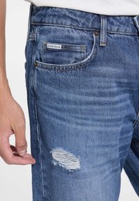 Vaqueros de mezclilla azul con un acabado desgastado, que presentan un pequeño detalle rasgado en el muslo y una etiqueta "GUESS JEANS" en el bolsillo trasero.
