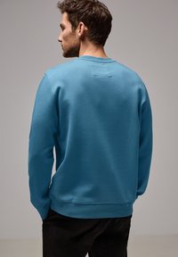 Hombre visto desde atrás, vestido con un sudadera azul simple y pantalones negros, de pie contra un fondo gris neutral.