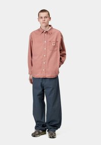 Camisa de mezclilla roja con botones, con un bolsillo en el pecho y detalle de logo, combinada con pantalones sueltos azul marino y zapatillas marrones con estampado.