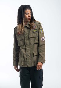 Oliwkowa kurtka wojskowa w stylu z licznymi kieszeniami i kolorowymi wyszywanymi naszywkami na ramionach. Wykonana z mocnego materiału, w luźnym kroju.