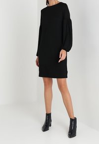 Vestido negro con mangas largas abullonadas, escote redondo y corte relajado. Estilizado con botas de tobillo negras brillantes. Textura de tela suave y lisa.
