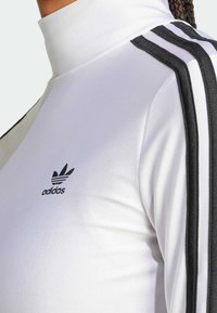 Vit långärmad tröja med hög krage, med svarta tre-rands accenter på axlarna och en liten svart Adidas-logotyp på bröstet.