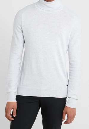 Person trägt einen hellgrauen Rollkragenpullover und schwarze Hosen, dargestellt vom Hals bis zur Hüfte vor einem einfarbigen Hintergrund.