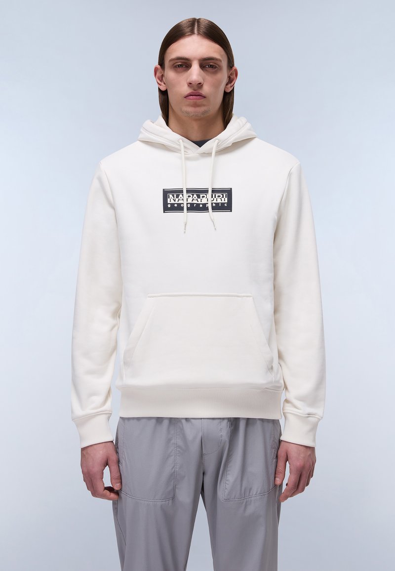 Bijela hoodie s prednjim džepom, kapuljačom sa šniranjem i crnim grafičkim logotipom. Obična tekstura tkanine, rebrasti rukavi i rub. Uparena sa sivim cargo hlačama.