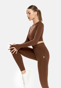 Barna, hosszú ujjú aktív viselet felső, bordázott textúrával, hozzá illő, testhez simuló leggings-szel párosítva. Logo díszítéssel rendelkezik. Fehér sportcipővel viselve.