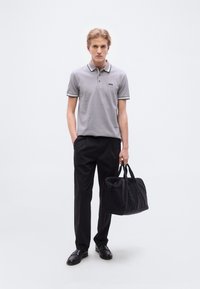 Γκρι πόλο μπλούζα με μαύρη επένδυση, λογότυπο στο στήθος, συνδυασμένη με μαύρο παντελόνι και παπούτσια. Το μοντέλο κρατά μια μαύρη τσάντα ταξιδιού, στέκεται μπροστά σε λευκό φόντο.