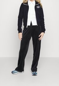 Černý sametový teplákový komplet s krátkým zipovým kabátem a volnými nohavicemi. Spárováno s modro-bílými sportovními botami. Pod tím bílá košile s knoflíky.