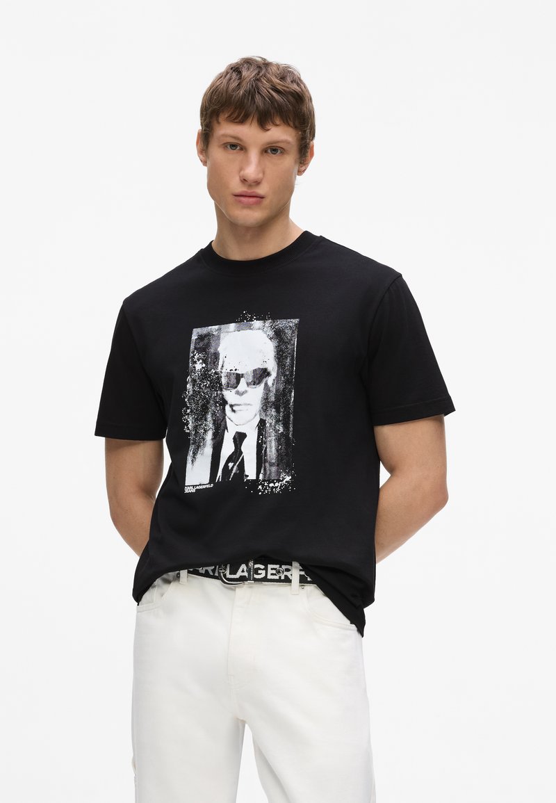 Νέος άνδρας φοράει μαύρο T-shirt με ασπρόμαυρο σχέδιο ενός άνδρα με γυαλιά ηλίου και κοστούμι, συνδυασμένο με λευκό παντελόνι.