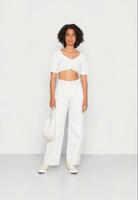 Blusa branca curta com mangas bufantes e um laço à frente, combinada com calças jeans brancas de cintura alta e uma bolsa de tecido trançado. Ténis brancos completam o look.