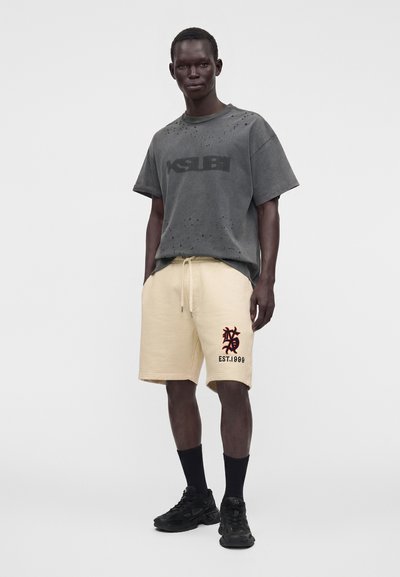 Homme debout portant un t-shirt noir délavé oversize, un short beige avec un emblème noir et rouge, des chaussettes noires et des baskets noires sur fond blanc.