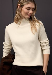 Kremowy sweter na drutach z wysokim prążkowanym kołnierzem i podwiniętymi mankietami, zestawiony z ciemnymi spodniami i brązową zamszową torbą.