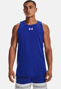 Mann mit Sommersprossen trägt ein ärmelloses blaues Under Armour-Shirt und blaue und weiße Sportshorts vor einem schlichten weißen Hintergrund.
