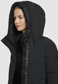 Sort pufferjakke med stor hætte, lynlås og trykknap lukninger samt strukturstof. Har læderdetaljer og et quiltet mønster.