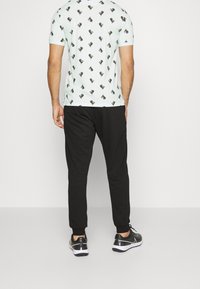 Hombre vestido con una camisa de color azul claro con un estampado de perros negros, pantalones de chándal negros y zapatillas negras, de pie de espaldas sobre un fondo sencillo.