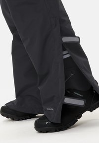 Des pantalons étanches noirs présentent des ouvertures ajustables, des accents en velcro gris et sont associés à des chaussures noires à forte adhérence.