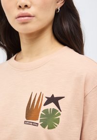 Κοντινό πλάνο μιας γυναίκας που φοράει μπεζ πουκάμισο με αφηρημένα σχέδια σε καφέ, μαύρα αστέρια και πράσινα φύλλα, καθώς και ένα μικρό λογότυπο "Scotch & Soda".