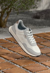 Sneaker bianca con accenti grigi e dettaglio del tallone marrone. Presenta una suola testurizzata e un design con lacci, posizionata su uno sfondo di piastrelle in legno e ghiaia.