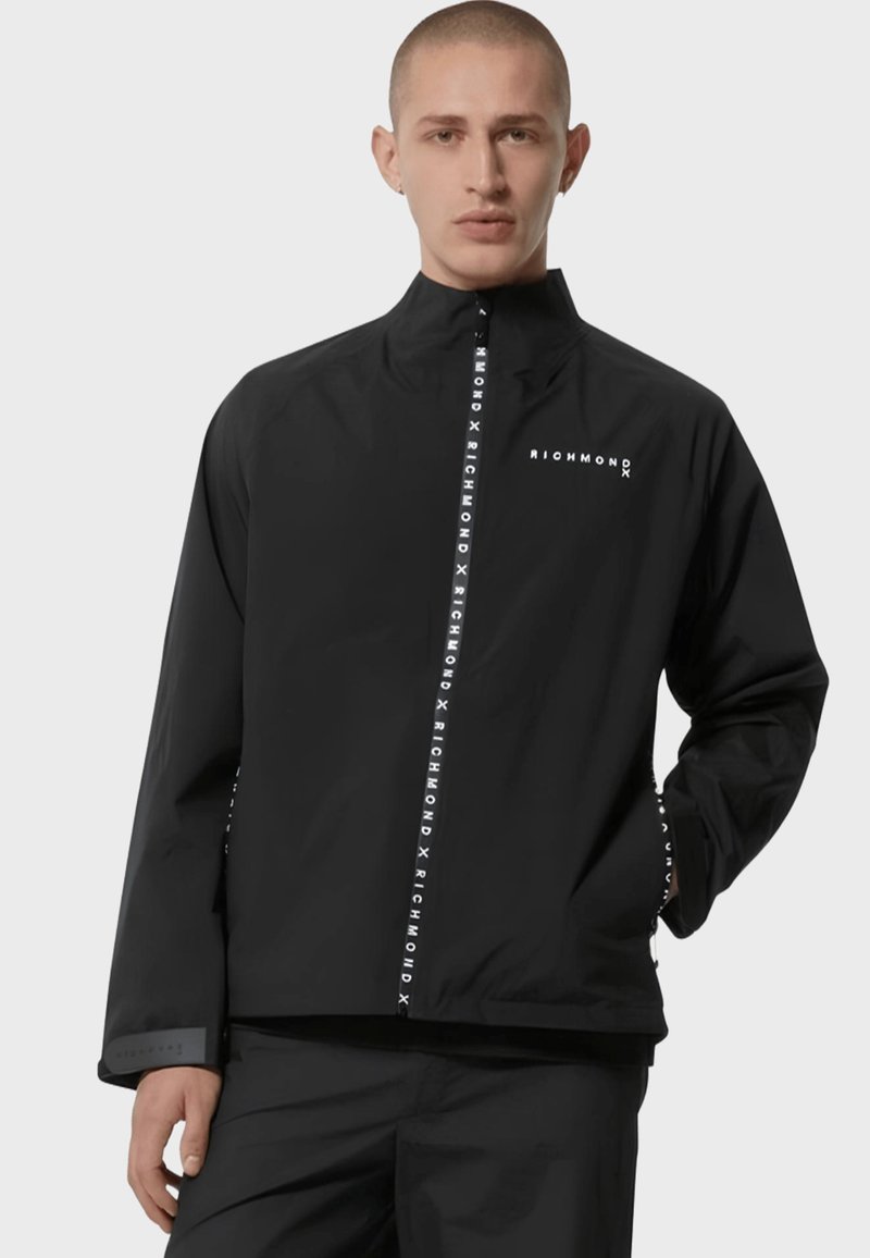 Giacca impermeabile nera con collo alto, chiusura a zip frontale e dettagli con logo sulla zip e sulle maniche. Il materiale appare liscio e leggero.