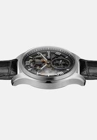 Skeleton horloge met een zilverkleurige roestvrijstalen kast, zwarte leren band met krokodillenstructuur, transparante wijzerplaat, Romeinse cijfers en gouden accenten.