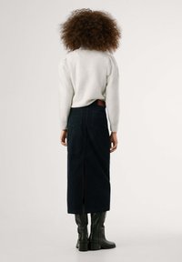 Hellgrauer Pullover mit pufferten Ärmeln, kombiniert mit einem marineblauen Cordrock mit einem Schlitz hinten und zwei Gesäßtaschen. Schwarze kniehohe Stiefel.