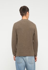 Brązowy sweter robiony na drutach z okrągłym dekoltem i długimi rękawami, charakteryzujący się teksturowanym wykończeniem i prążkowanym dołem, noszony z jasnoniebieskimi dżinsami.