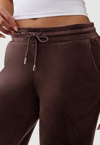 Środkowa część ciała osoby w brązowych welurowych dresowych spodniach z wiązaniem w talii oraz białym krótkim topie, ręka dostosowująca pas.