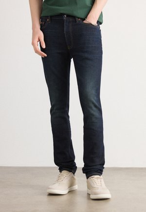 Dark blue slim-fit jeans with light fading, featuring five pockets and a mid-rise. Paired with beige textured sneakers.