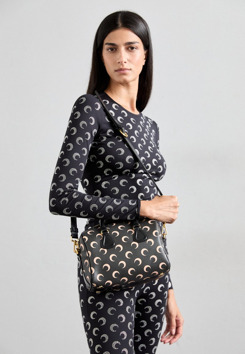 Black leather handbag with a pastel crescent moon pattern, dual top handles, and an adjustable strap, complementing a patterned black outfit.