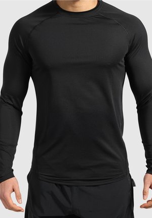Homme portant un haut de sport noir moulant à manches longues et un pantalon noir, debout avec les bras détendus le long du corps.