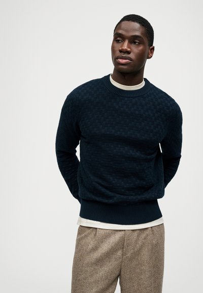 Dunkelblauer Pullover mit strukturiertem, gewebtem Muster und gerippten Bündchen sowie Saum. Kombiniert mit einem hellen Hemd und braunen, gemusterten Hosen.