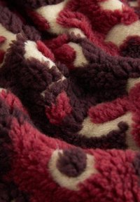 Tkanina o fakturze w kolorach czerwonym, bordowym i kremowym z wijącym się wzorem. Materiał wydaje się miękki i puszysty, o grubym, puszystym włóknie.